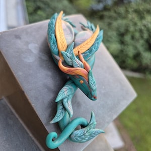 Könnte beinhalten: Eine modellierte Drachenfigur mit blaugrauen, blattartigen Schuppen, orangefarbenen und gelben Hornakzenten sowie zwei glatten, abgerundeten Steinen auf dem Rücken. Das Wesen hat einen geschwungenen, schlangenförmigen Körper mit geringeltem Schwanz.