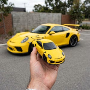 Può includere: Un modellino di auto sportiva giallo con ruote nere e alettone posteriore, tenuto in primo piano, con un'auto sportiva gialla a grandezza naturale con design abbinato e grande alettone posteriore visibile sullo sfondo sfocato.