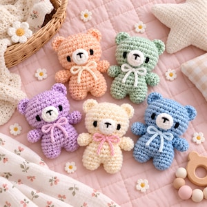 Könnte beinhalten: Fünf kleine gehäkelte Teddybären in Pfirsich, Mintgrün, Lavendel, Creme und Blau. Jeder Bär hat eine weiße Schnauze, schwarze Augen und eine kleine Schleife am Hals. Die Bären sind auf einer gesteppten rosa Oberfläche mit kleinen weißen Blumen angeordnet.