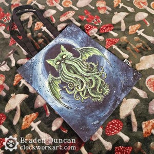 Peut inclure: Un sac fourre-tout noir avec une illustration colorée d'une créature ressemblant à un chat avec des tentacules. La créature est verte et violette et a un fond étoilé. Le sac porte le texte "Braden Duncan clockworkart.com" imprimé en bas.