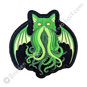 May include: Green embroidered patch of a cat with a tentacled beard and wings. The cat has yellow eyes and a black background.
