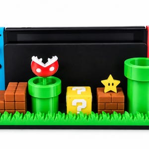 Può includere: Una base di ricarica per console di gioco nera a tema Super Mario, dotata di tubi verdi, scale in mattoni marroni, un blocco punto interrogativo giallo, una stella gialla e una pianta piranha rossa e bianca su una piattaforma d'erba verde.
