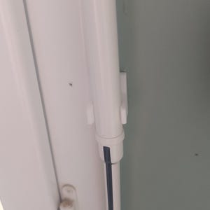 Op de afbeelding: Een witte kunststof lamellendrager voor verticale jaloezieën bevestigd aan een witte verticale rail. Het mechanisme heeft een cilindrische behuizing met een kleine uitstekende clip en een dunne zwarte lijn op de onderste stang.