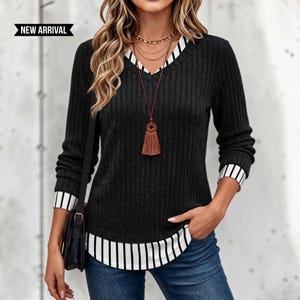 May include: A charcoal gray ribbed long-sleeve sweater featuring a V-neckline with a white and black vertical striped trim, matching striped cuffs, and a striped hemline. The garment includes a black bag and a brown tassel necklace. Text reads NEW ARRIVAL.