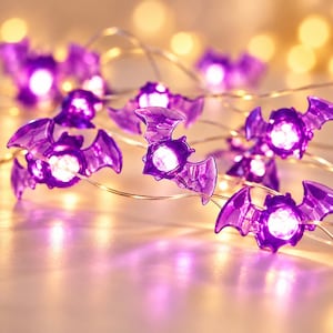 Può includere: Una catena di luci decorative con coperture traslucide a forma di pipistrello di colore viola. Ogni pipistrello contiene una piccola lampadina LED bianca brillante che si illumina dall'interno. Le figure sono collegate da un filo sottile e flessibile.