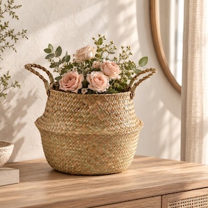 May include: A woven seagrass basket with two braided handles, featuring a tapered shape and a natural tan and light green colour palette. The basket is filled with a bouquet of soft pink roses, small white filler flowers, and green eucalyptus leaves.