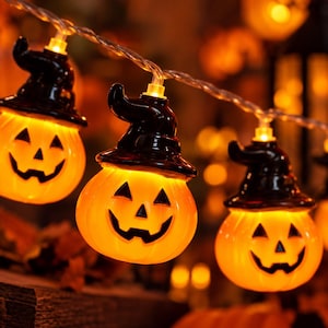 Può includere: Una catena di luci decorative con zucche arancioni che indossano cappelli da strega neri. Ogni zucca presenta un volto intagliato con occhi triangolari e una bocca frastagliata, illuminata dall'interno da una luce calda collegata da un filo.