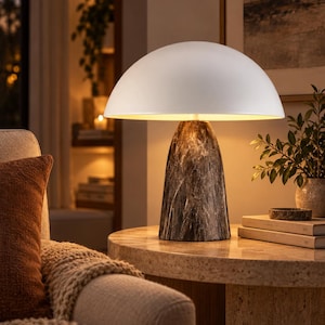 May include: A table lamp featuring a wide, smooth, matte white dome-shaped shade positioned atop a conical base with a dark, marbled pattern. The lamp emits a warm, soft glow from beneath the shade, illuminating the surface it rests upon.