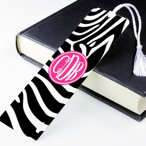 May include: A black and white zebra print bookmark with a pink circle containing the monogram "CDB". The bookmark has a white tassel.