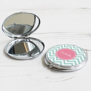 May include: A silver compact mirror with a green and white chevron pattern on the front. The mirror has a pink circle with the name "Isabella" written in white.