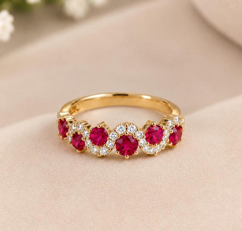 May include: A gold band ring featuring a wavy design set with five round-cut vibrant red stones and small clear white stones arranged in a delicate pattern along the top half of the band, creating a continuous sparkling effect across the metal surface.