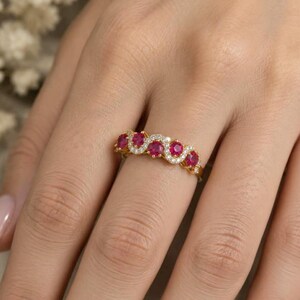May include: A gold ring featuring four round, vibrant red gemstones set in a row, accented by a delicate border of tiny, sparkling white stones that follow the wavy, scalloped edge of the band, creating a detailed and elegant jewellery piece.