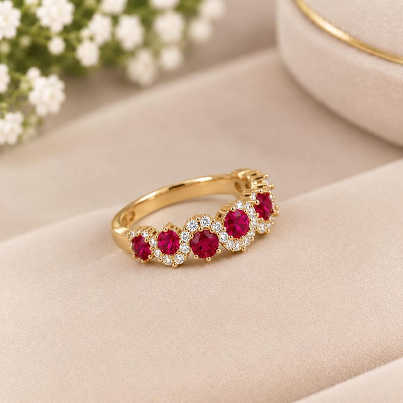 May include: A gold ring featuring a curved, wave-like band set with five round, vibrant red gemstones. Each red stone is surrounded by a halo of small, clear, sparkling white stones, creating a continuous, elegant, and detailed jewellery piece.