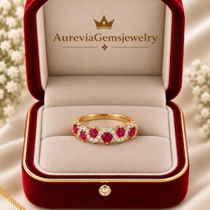 May include: A gold ring featuring a wavy band set with round red rubies and small white diamonds, presented in a red velvet box with the text AureviaGemsjewelry printed in gold on the inner lid. The ring sits centred on a beige velvet cushion.