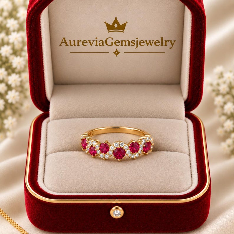 May include: A gold ring featuring a wavy band set with round red rubies and small white diamonds, presented in a red velvet box with the text AureviaGemsjewelry printed in gold on the inner lid. The ring sits centred on a beige velvet cushion.
