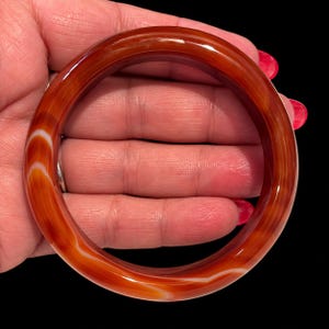 May include: A smooth, polished bangle bracelet crafted from translucent orange and red carnelian stone. The circular jewelry piece features distinct, swirling white banded patterns throughout the material, creating a natural, layered appearance with a glossy finish.