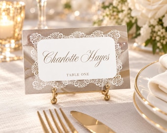 Op de afbeelding: Een rechthoekig naamkaartje met een wit middenpaneel met de gedrukte naam Charlotte Hayes en de tekst TABLE ONE, omringd door een witte kanten rand op een taupekleurige achtergrond, geplaatst op een kleine gouden standaard.