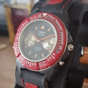 May include: A Sector chronograph wristwatch featuring a black dial with two sub-dials, a red rotating bezel with white numerals, and a black fabric strap with red stitching. The watch face displays the text Sector, Undersea, and 100 Meters.
