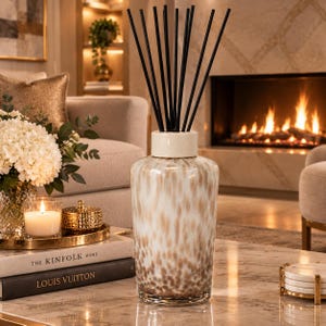 May include: A glass reed diffuser with a speckled white and brown pattern, featuring a cylindrical body and a solid white neck. The diffuser is topped with a set of thin, straight black reeds extending upwards from the centre of the vessel.