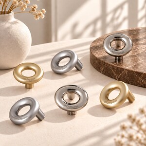Può includere: Una collezione di cinque pomelli circolari per armadietti con finiture in oro spazzolato e argento lucido. Ogni pomello presenta un design ad anello con un'apertura centrale e il testo "zusaptoOftc" inciso sulla superficie, montato su una base cilindrica.