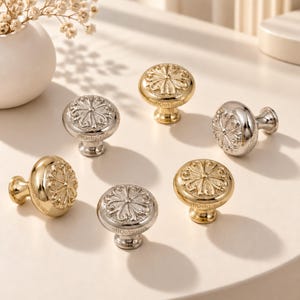 May include: A collection of six round cabinet knobs in gold and silver metallic finishes. Each knob features a raised, intricate floral-inspired pattern on the circular face and a smooth, tapered base for mounting to furniture or cabinetry.