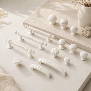 May include: A collection of matte white cabinet hardware including three long cylindrical pulls with ribbed ends, two arched pulls, and several round knobs in smooth, fluted, and tiered designs. Each piece features the text "zuaploOffc" printed on it.