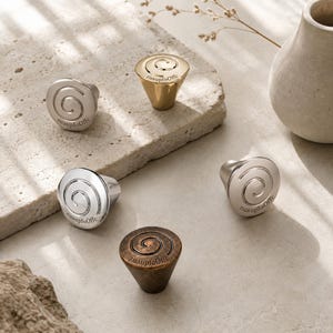 May include: Five metal cabinet knobs in various finishes including gold, silver, chrome, and bronze. Each knob features a circular top with a spiral design and the text zuaupfoOffc engraved on the surface. The knobs have a tapered base design.