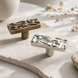 May include: Two rectangular cabinet knobs with an irregular, textured surface. One knob features a matte antique brass finish, while the other has a polished silver-tone finish. Both knobs are engraved with the text zuaupfoOffc on the front.