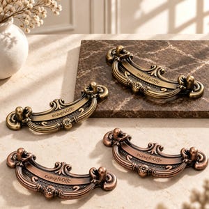 Puede incluir: Cuatro tiradores de cajón de metal curvos y ornamentados con intrincados adornos y un motivo floral central. Dos tiradores tienen un acabado de latón cepillado y dos tienen un acabado de cobre. Cada tirador tiene grabado el texto zuaupfoOffc.