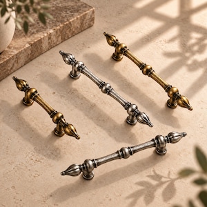 May include: Four metal cabinet pulls arranged in a group. Two pulls feature a brushed brass finish, while the other two have a weathered silver finish. Each pull has a central bar with decorative rounded segments and pointed finial ends.