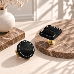 Puede incluir: Dos pomos para armarios con tapas de cuero negro, costuras blancas en contraste y bases de metal dorado. Un pomo es cuadrado y el otro es circular. Ambas piezas tienen el texto "XianpfoOfe" grabado en la superficie negra.