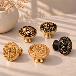 Könnte beinhalten: Fünf runde Schrankknöpfe mit komplexen Prägemustern. Vier Knöpfe haben ein metallisches Gold-Finish mit verschiedenen Blumen- und Geometriemustern, während ein Knopf eine schwarze Basis mit goldenen Blumendetails auf der Vorderseite aufweist.