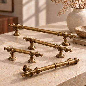 May include: Four antique brass cabinet pulls of varying lengths, each featuring a cylindrical bar with decorative finials and two mounting posts. The metal has a brushed, weathered finish with the brand name "ZumpOtte" engraved on the bars.