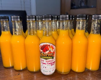 May include: A collection of clear glass bottles filled with bright orange hot sauce, each topped with a black cap. The front-most bottle features a label with a red horse graphic, the text Burns Peppers Hot Sauce Cosmic Cantaloupe, and a best by date of 9/27.