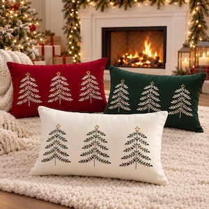 Puede incluir: Tres cojines rectangulares en rojo, verde oscuro y crema. Cada cojín presenta un diseño bordado de tres árboles de Navidad estilizados, cada uno rematado con una estrella dorada y decorado con pequeños detalles coloridos.