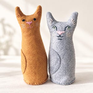 Puede incluir: Dos figuras de gato de fieltro hechas a mano, una en color marrón dorado cálido y la otra en gris claro suave. Cada gato presenta detalles faciales bordados, incluyendo una nariz rosa cosida, ojos oscuros y bigotes blancos, con un cuerpo redondeado.