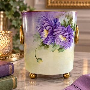 Puede incluir: Un recipiente cilíndrico de cerámica con un diseño pintado a mano de grandes crisantemos morados sobre un fondo degradado de amarillo pálido y lavanda. La pieza incluye dos asas laterales doradas y tres pequeños pies dorados.