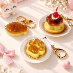 May include: Three keychains featuring miniature food-themed charms: a brown fish-shaped cake, a yellow custard pudding topped with white cream and a red cherry on a white plate, and a yellow paw-shaped jelly on a white plate, all with gold rings.