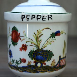 May include: A white ceramic canister with a lid, featuring the word PEPPER in black lettering. The exterior is decorated with hand-painted floral motifs in red, blue, green, and yellow, with a green rim on the lid and base.