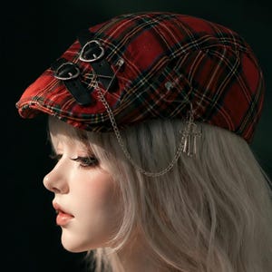 Puede incluir: Una gorra plana de cuadros rojos y negros con dos correas de cuero negro con hebillas plateadas en la parte delantera, un imperdible plateado y una cadena plateada con un dije de cruz colgando del lateral. El texto PeachybyRyn es visible.
