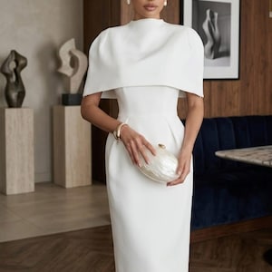 May include: A white midi dress featuring a high neckline and a structured cape-style overlay that covers the shoulders. The dress has a fitted waist and a straight skirt silhouette. A small, oval-shaped, iridescent white clutch is held in the center.