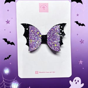 May include: A hair bow shaped like bat wings, featuring a black glitter base and two purple glitter sections. The bow is attached to a white display card with the text @beautiful bows and clips printed in small black letters at the bottom.
