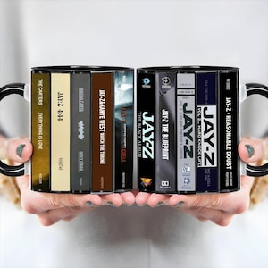 May include: A ceramic mug featuring a wrap-around graphic of various Jay-Z cassette tape spines, including titles like The Black Album, The Blueprint, Reasonable Doubt, 4:44, and Watch The Throne, with black handles on both sides.