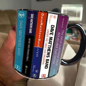 May include: A ceramic mug featuring a graphic print of five Dave Matthews Band cassette tape spines. The designs include Everyday, Before These Crowded Streets, Crash, Under the Table and Dreaming, and Remember Two Things with visible RCA and BNR logos.