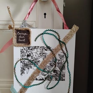 May include: A rectangular white wall hanging featuring a black and white floral illustration, a small square sign with the text "Courage, dear heart," a pink ribbon hanger, gold glitter trim, and decorative teal and silver ribbon accents.