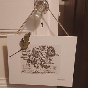 Könnte beinhalten: Ein rechteckiges weißes Schild, das an einem klaren Glas-Türknauf mit einer Juteschnur hängt. Das Schild zeigt eine schwarz-weiße Blumenillustration, kursiven Text und ein kleines getrocknetes grünes Blatt in der oberen linken Ecke.