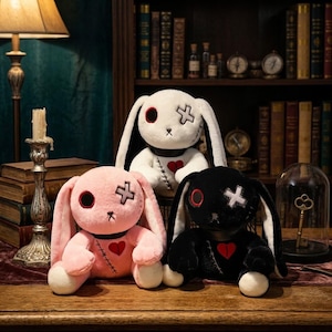May include: Three plush rabbit toys in white, pink, and black. Each rabbit features long ears, a circular eye, an X-shaped eye patch, a stitched chest detail, and a red heart appliqué. The toys are arranged in a group on a wooden surface.