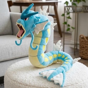 May include: A blue and yellow plush toy shaped like a sea serpent with a wide open mouth, sharp white teeth, red eyes, and white fins along its back and tail. The creature has a long, curved body with yellow oval markings and blue whiskers.