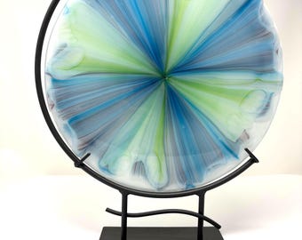 May include: A decorative glass plate with a radial design in shades of blue, green, and white, displayed on a black metal stand. The plate's design resembles a flower or burst of color. The stand has a square base and a curved support.