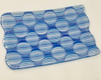 May include: A blue and white glass dish with a wavy, striped pattern. The dish has a slightly rounded rectangular shape with a wavy edge.